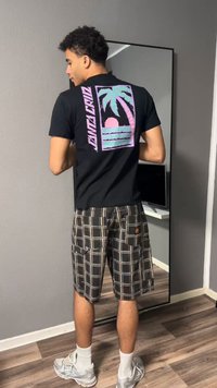 Νέος άνδρας που φοράει μαύρο T-shirt με παστέλ γραφικό με φοίνικα και καρό cargo σορτς, στέκεται μπροστά σε καθρέφτη μέσα στον χώρο.