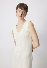 Úzky bezrukávový šaty v svetlej krémovej farbe s vertikálnou ryhou a výstrihom do V. Hladká tkanina bez viditeľných zapínaní.