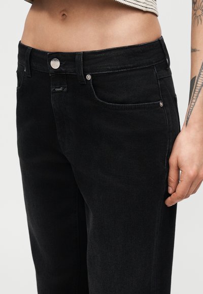 Gros plan sur une personne portant un jean noir taille haute avec un bouton argenté et un haut court à rayures, montrant un bras tatoué.