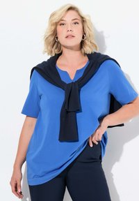 Modrá košile s krátkým rukávem a výstřihem do V, doplněná tmavě modrým svetr na krku. Hladká textilie. Tmavé kalhoty dotvářejí outfit.