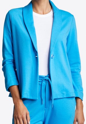 Persoon in een helderblauwe blazer met één knoop, over een witte top en bijpassende blauwe sweatpants met koordjes, staand tegen een witte achtergrond.