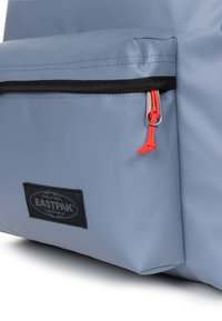 Sac à dos Eastpak gris avec une poche frontale zippée, fermoir noir, tirette rouge et un patch logo tissé en bas. Matériau texturé.