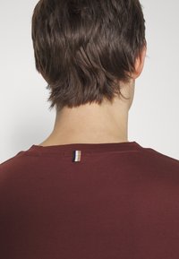 Burgundské tričko s kulatým výstřihem, krátkým rukávem a malou pruhovanou nášivkou na zadní straně poblíž výstřihu. Měkká textura tkaniny.