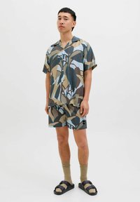 Kortærmet skjorte med knapper og bladmønster i jordfarver; matchende shorts; båret med brune sokker og sorte sandaler.