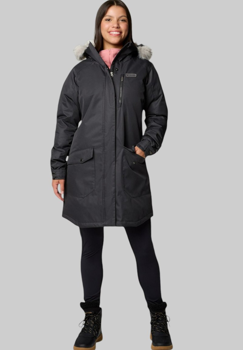 Schwarze Parka-Jacke mit einer mit Fell gefütterten Kapuze, ausgestattet mit einem Reißverschluss vorne, zwei großen Fronttaschen und einem mittellangen Design. Getragen über schwarzen Leggings.