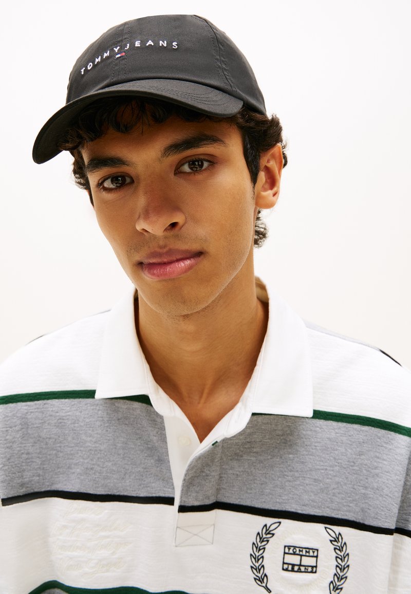 Μαύρο καπέλο με κέντημα "TOMMY JEANS"; ριγέ πόλο μπλούζα σε λευκό, γκρι και πράσινο με λογότυπο και διακοσμητικό κέντημα στο στήθος.