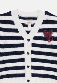 Ριγέ γιλέκο με ναυτικό και λευκό χρώμα, ραβδωτή υφή, V-γυ neckline και κουμπιά σε σχήμα καρδιάς. Εντυπωσιακή λεπτομέρεια καρδιάς από κόκκινα πούλια.