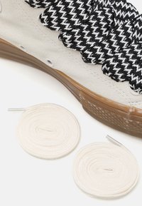 Witte sneaker met een gestructureerde bovenkant en geweven zwart-witte veters. Inclusief twee opgerolde, witte katoenen veters met zilveren aglets.