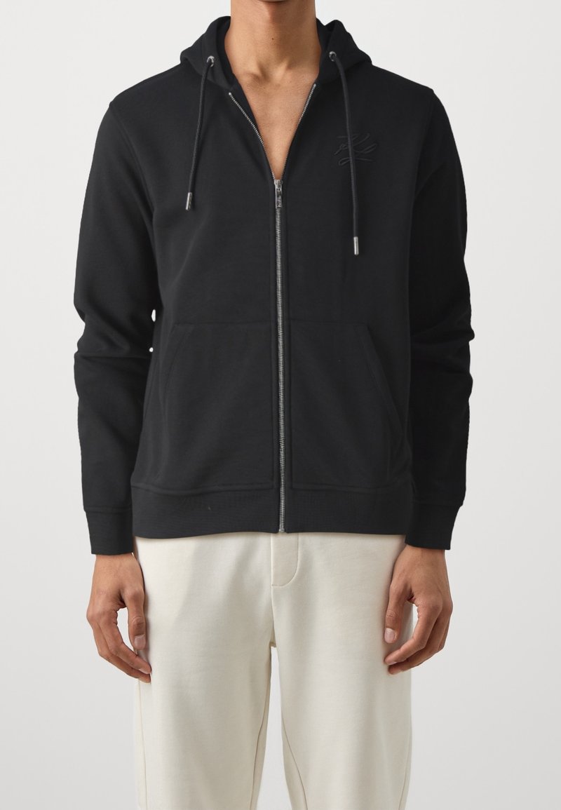Černá mikina na zip s klokaní kapsou a kapucí se šňůrkou. Obsahuje texturované logo na hrudi a žebrované manžety. Měkký materiál.