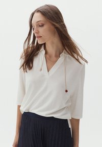 Blusa bianca a maniche corte con scollo a V e texture morbida, abbinata a una gonna scura a fantasia. Gioielli semplici accentuano l'outfit.