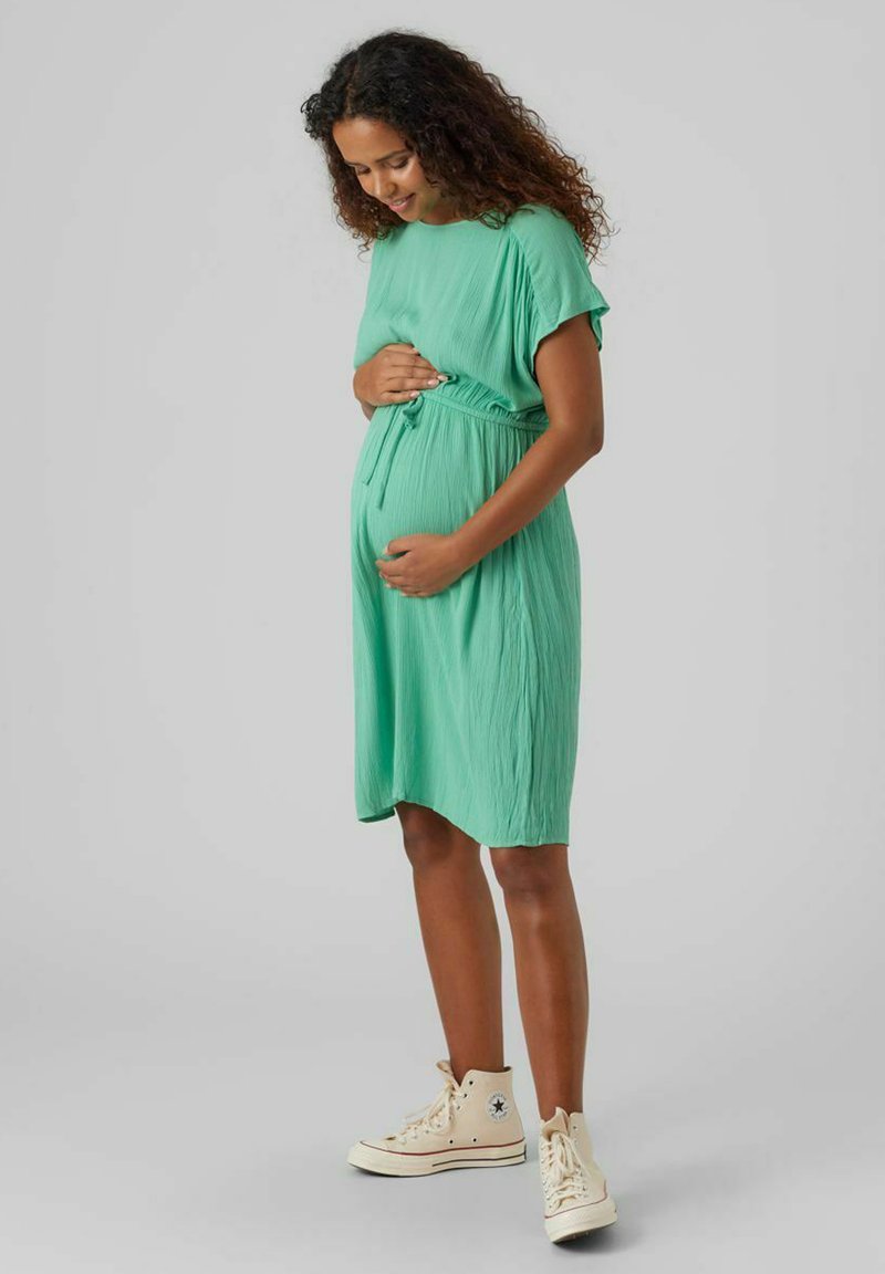 Robe de maternité plissée verte avec des manches courtes, taille cintrée et coupe décontractée, associée à des baskets montantes beiges.