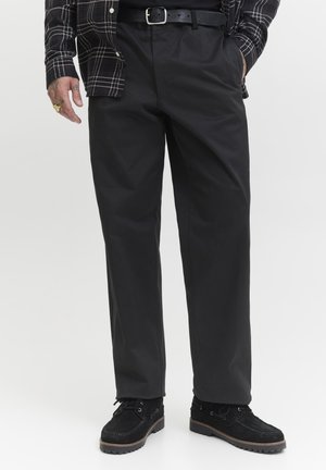 Hombre con pantalones negros de pierna recta, cinturón negro, zapatos de ante negros y una camisa de cuadros en blanco y negro con las mangas abajo.