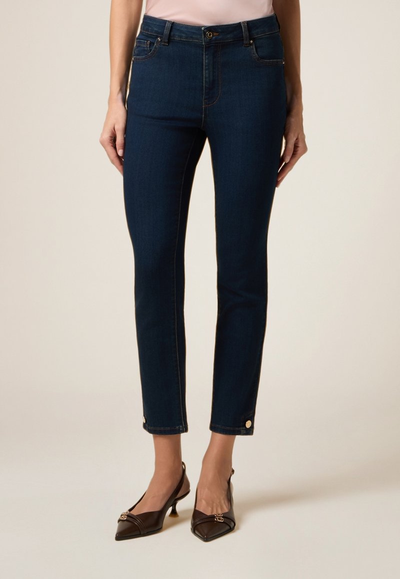 Jean slim fit bleu foncé en denim à la texture lisse. Comprend des poches avant et des accents de boutons dorés à l'ourlet.