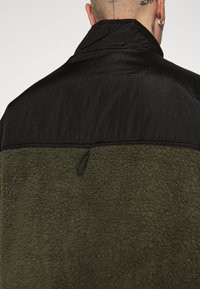 Fleece jack met een zwarte, gladde topsectie en een olijfgroene, gestructureerde ondersectie; bevat een lusdetail aan de achterkant.