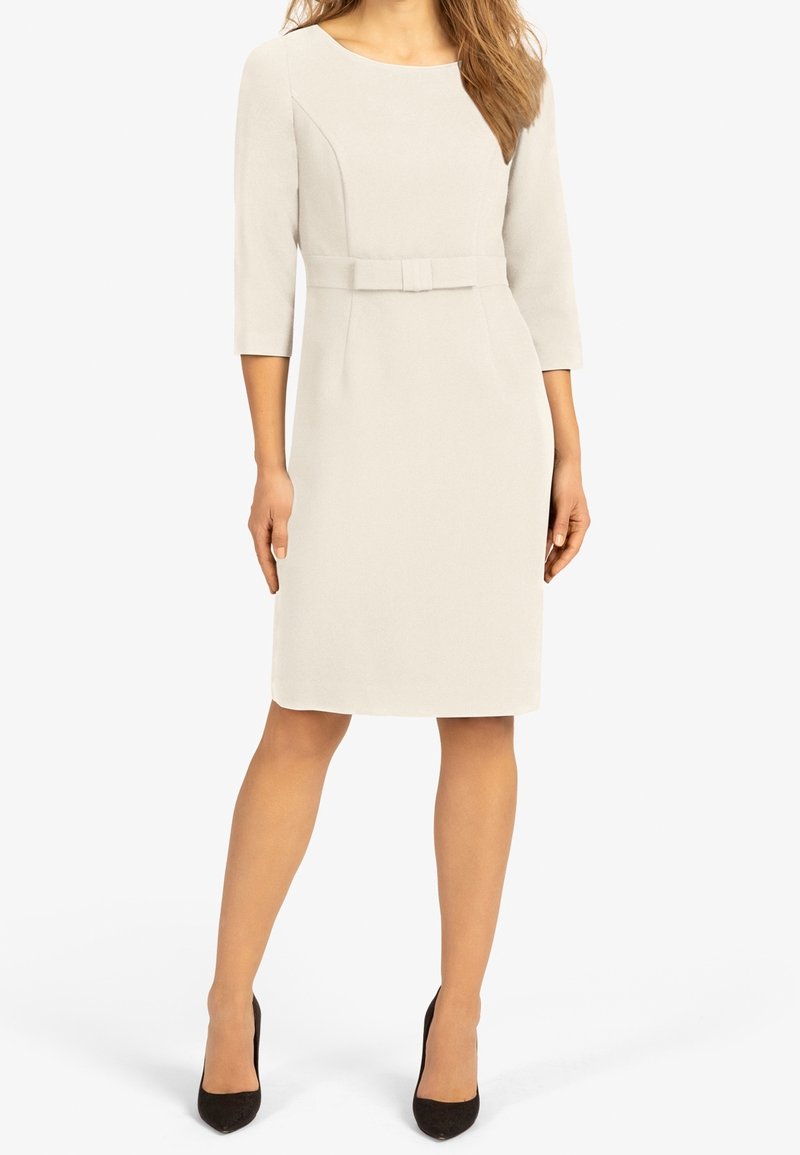 Une robe couleur crème à mi-genou avec des manches trois-quarts, un col arrondi et une taille cintrée mise en valeur par une ceinture en tissu.
