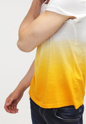 Personne portant un t-shirt à manches courtes dégradé jaune et blanc avec un jean bleu foncé, bras plié et main proche du visage.
