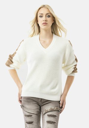 Witte gebreide truien met een diepe V-hals en gek structureerde mouwen in bruin. Het model draagt verouderde lichtgrijze jeans met scheuren en gestikte accenten.