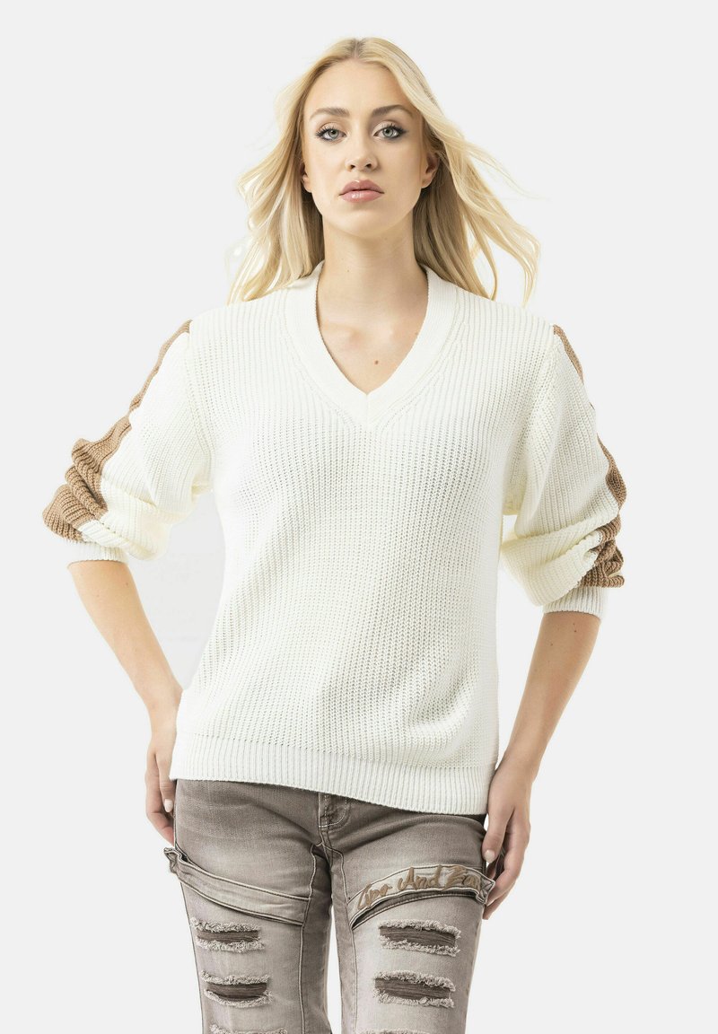 Witte gebreide truien met een diepe V-hals en gek structureerde mouwen in bruin. Het model draagt verouderde lichtgrijze jeans met scheuren en gestikte accenten.