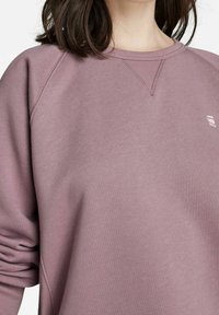 Žena v mauve svetru s kulatým výstřihem, na levém prsu s malým bílým logem, má dlouhé tmavé vlasy, které jsou částečně vidět.