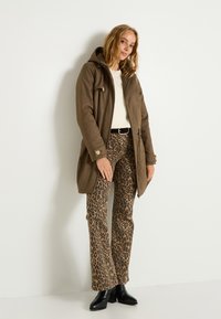 Parka marrón con capucha, cuenta con cierre de cremallera y botón. Pantalones de campana con estampado de leopardo, botas negras hasta el tobillo y una blusa crema completan el look.
