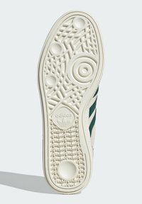 Biała podeszwa sneakerów z teksturowanymi wzorami, zawierająca okrągłe i liniowe desenie. Na boku widoczne są zielone paski akcentujące.