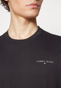 Camiseta de algodón negra con cuello redondo, que presenta un logo blanco de "TOMMY JEANS" y un pequeño emblema de colores en el lado izquierdo del pecho. Textura suave.