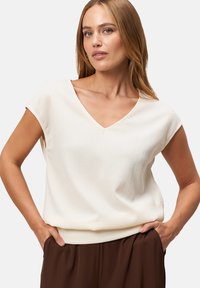 Light beige short-sleeve top with a V-neck and textured surface; worn with brown pants, featuring a relaxed fit and soft fabric.