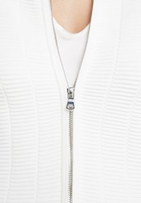 Biała kardiganka na zamek z teksturowanymi paskami, z metalowym zamkiem błyskawicznym. Tkanina wydaje się gładka i lekko elastyczna, nałożona na biały top.