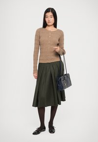 Brown ribbed cardigan with buttons, green pleated skirt, black tights, and black flats, holding a navy handbag with a buckle detail.