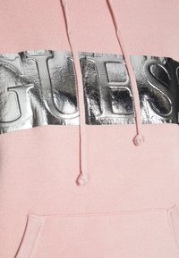 Felpa rosa con cappuccio, caratterizzata da un logo "GUESS" metallico argento sul petto, con cordini e tasca frontale. Tessuto morbido.