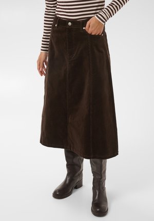 Mujer con falda midi marrón oscuro, camiseta de manga larga a rayas y botas de cuero marrón oscuro hasta la rodilla, de pie con una mano en el bolsillo.