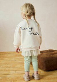 Kermainen collegepaita, jossa on tummansininen "Dancing Friends" -teksti, kerroksellinen tyllyhame, mintunvihreät ribbihousut ja beige nilkkasaappaat. Puulattian tausta.