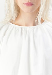 Hvit bluse med løs passform, rynket halsutringning og myk tekstur. Enkelt design med minimale synlige detaljer.