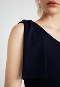 Tamsiai mėlyna suknelė su drapiruota peties detale, pagaminta iš lygaus audinio, turinti gilia V formos iškirptę ir laisvai prigludusį siluetą.