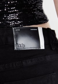 Tela de denim negro con una etiqueta de cuero brillante que muestra el nombre de la marca "l.t.b" y el texto "LIVE TO BE" y "STUDIO DESIGNED" embosados en blanco.