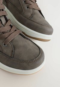 Bruna texturerade sneakers med stadiga snören, med en vit mellansula och en gummibelagd yttersula. Närbild visar sömdetaljer.