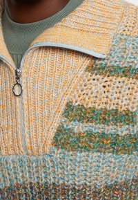 Zbliżenie na wielokolorowy sweter dziergany w żółte, zielone i niebieskie wzory oraz srebrny zamek błyskawiczny z okrągłym uchwytem.