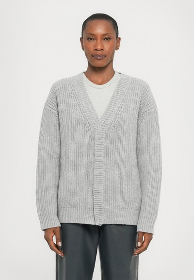 Cardigan tricoté gris avec un col en V et une ouverture devant. Présente des poignets et un ourlet côtelés. Tissu texturé avec une coupe ample. Porté sur un haut gris clair.