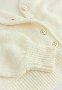 Cardigan en crème fabriqué à partir d'un tissu doux et texturé. Présente des poignets côtelés et des boutons dorés en forme de cœur avec des accents décoratifs.