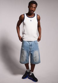 Weißes, geripptes Tanktop mit blauen Akzenten, kombiniert mit hellblauen, ausgefransten Jeansshorts. Schwarze Sneaker mit blauen Details.
