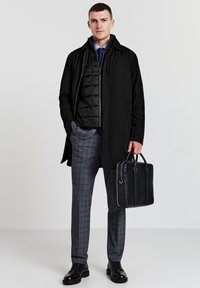 Schwarzer langer Mantel über einer gesteppte Weste, graue karierte Hose und schwarze Schuhe. Hält eine schwarze Ledertasche. Einfaches und modernes Design.
