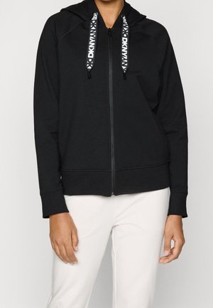 Persoon draagt een zwarte hoodie met rits met witte DKNY-logo koordjes en gebroken witte broek tegen een effen lichte achtergrond.