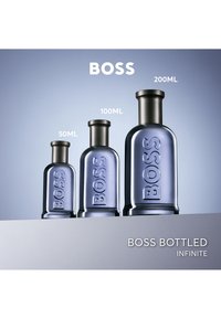Drie blauw getinte flesjes Boss Bottled Infinite parfum in 50 ml, 100 ml en 200 ml maten op een grijs oppervlak met een verloop van blauwe achtergrond.