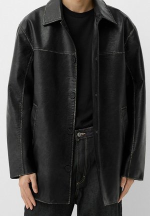 Veste en cuir noire à boutons portée sur une chemise noire avec un jean en denim foncé sur une personne debout devant un fond uni.