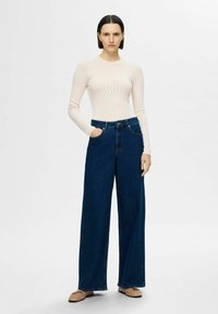 Stickad långärmad topp med ett texturerat zigzagmönster i ljusbeige, kombinerad med vida mörkblå denimjeans som har sidofickor.