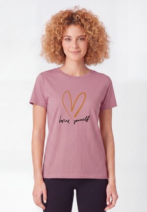 Femme aux cheveux bouclés portant un t-shirt mauve avec un cœur orange et le texte "love yourself", debout devant un fond blanc uni.