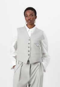 Gilet en tissu gris au design structuré, avec un devant à cinq boutons, deux poches latérales et un détail de poche carrée. Porté par-dessus une chemise blanche.