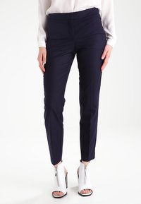 Marinblå slim-fit byxor med en mjuk textur och medelhög midja. Matchas med vita öppna högklackade skor med blixtlås.