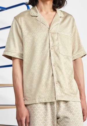 Personne portant une chemise de pyjama beige à manches courtes boutonnée et un short assorti avec un motif subtil de feuilles et un détail de passepoil blanc.