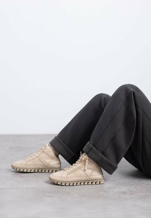 Zapatillas altas beige de cuero texturizado, con suela en zigzag y detalles de cremallera lateral, combinadas con pantalones negros de dobladillo enrollado.